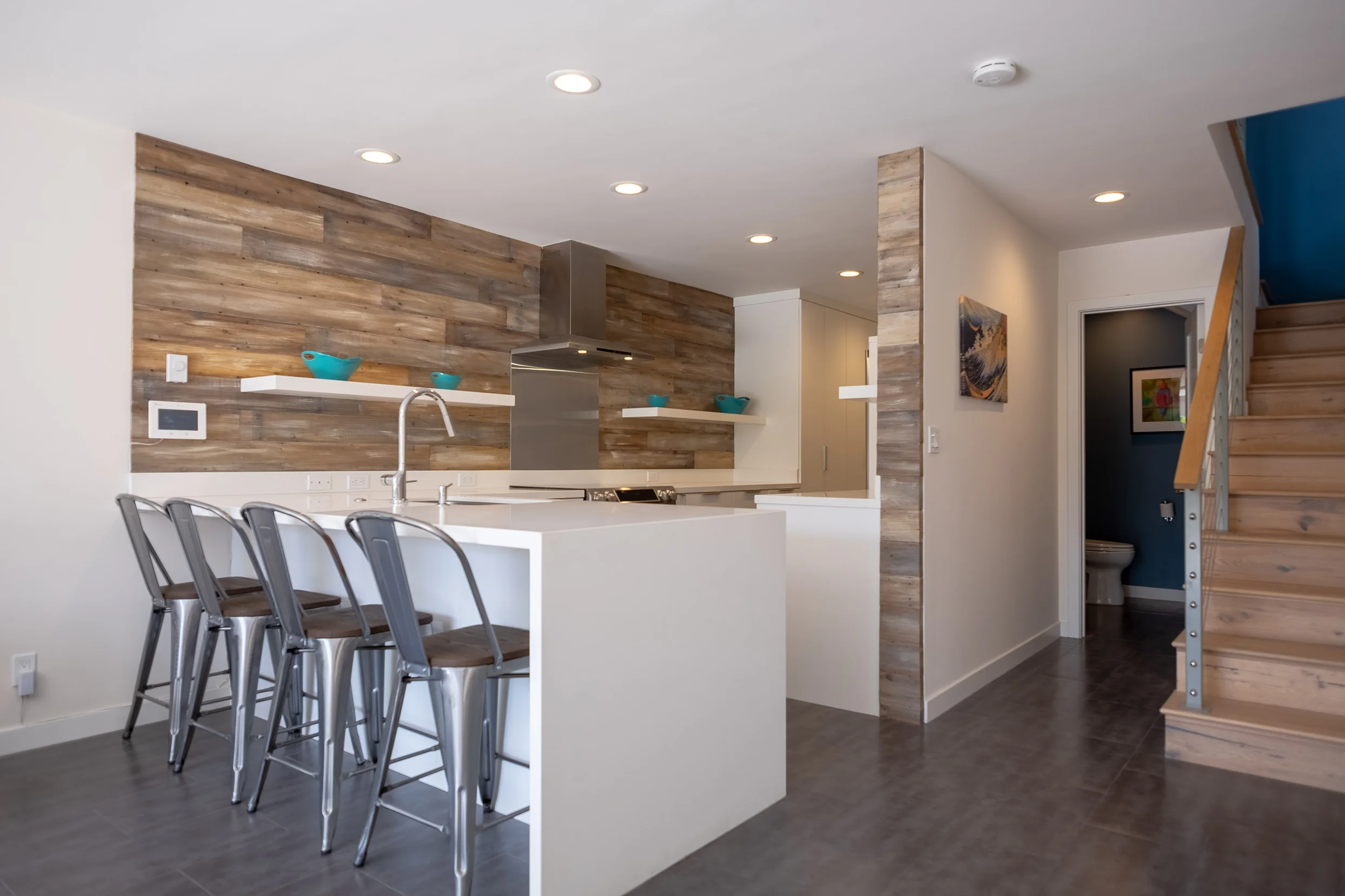 New kitchen interior with white quartz waterfall counter, white cabinets and shelves contrasting with grey tile, whitewashed redwood backsplash, oak stairs and blue accent walls.